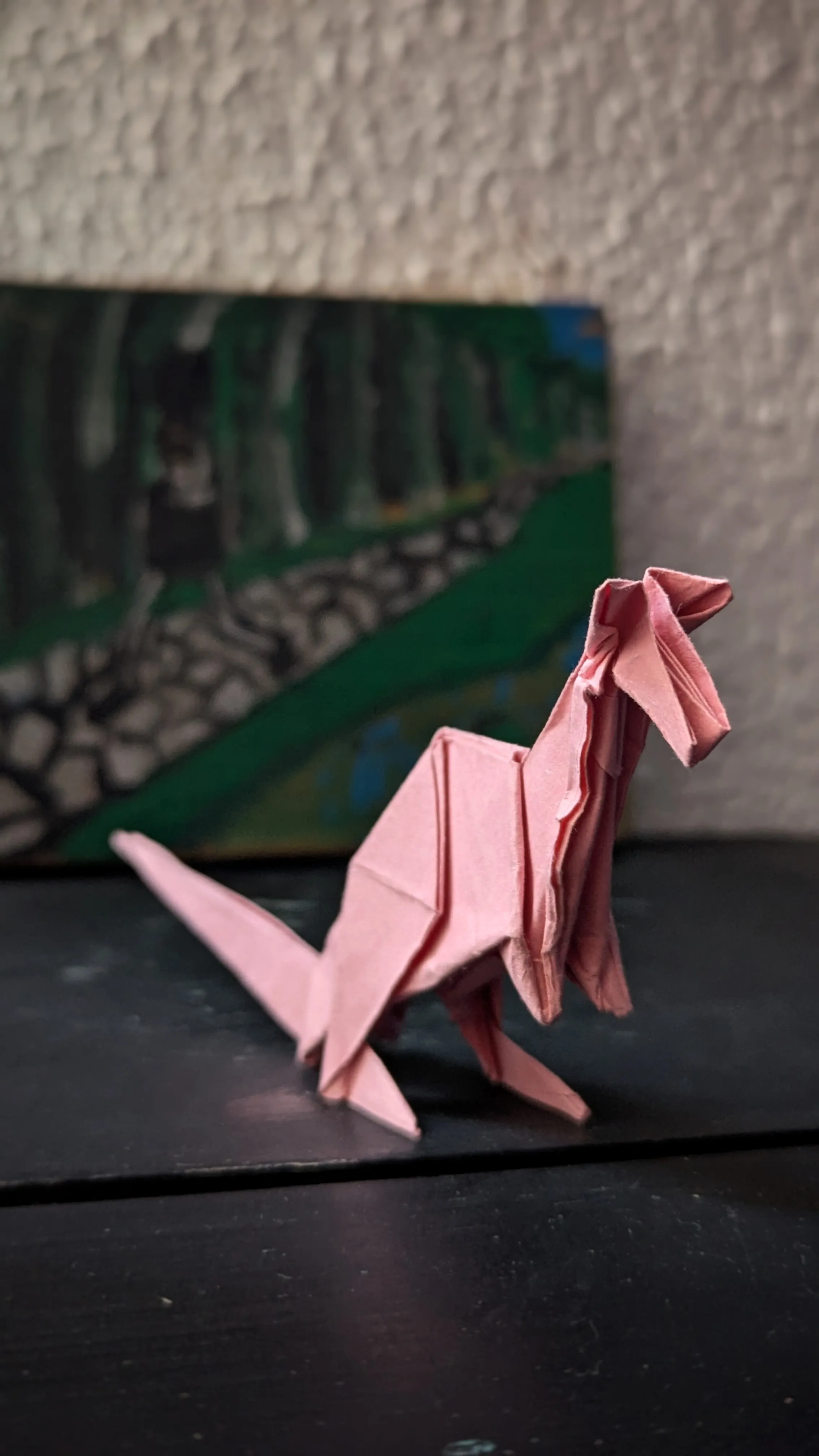 Un canguro de papel rosa apoyado en la cola sobre una mesa negra, con un cuadro verde apoyado en la pared detrás.