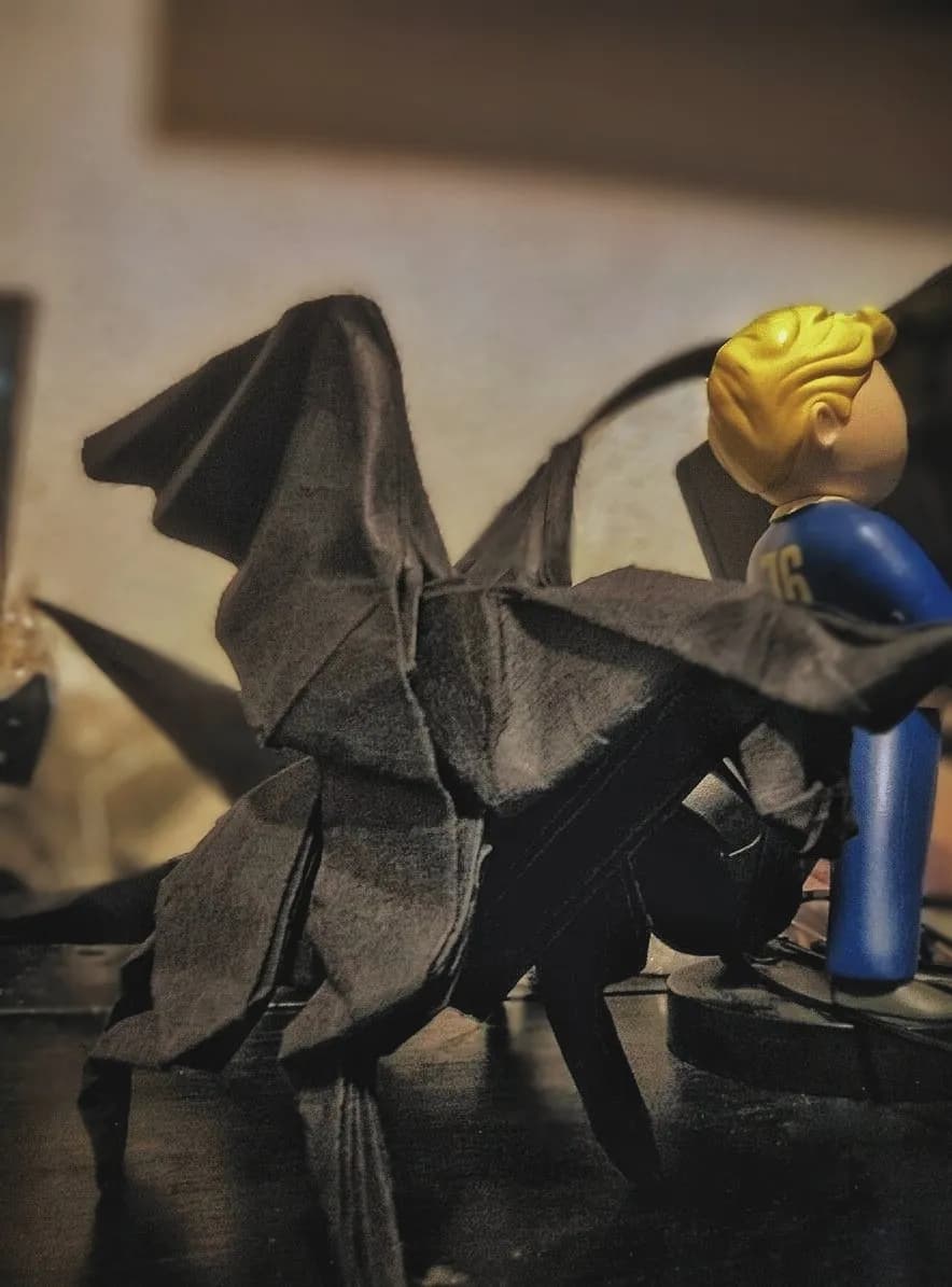 Un pegaso de papel gris oscuro con las alas desplegadas sobre un estante, junto a un muñeco rubio con traje azul.