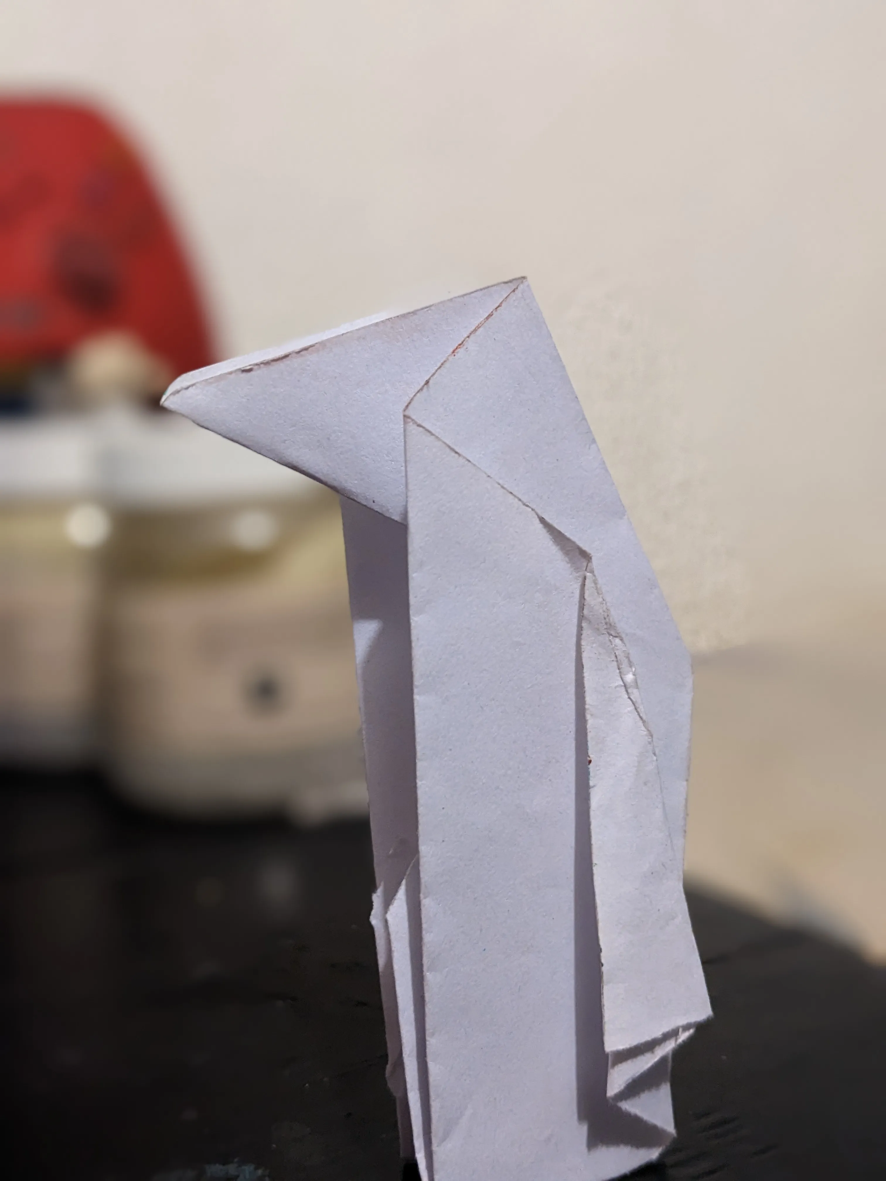 Un pingüino de papel blanco de pie sobre una mesa oscura, contra una pared blanca, con un objeto rojo desenfocado a un lado.